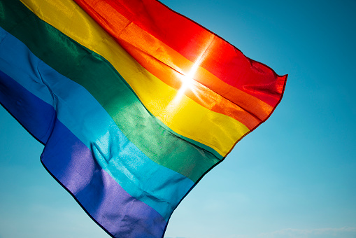 closeup of a rainbow flag waving on the blue sky, moved by the wind, with the sun in the background
