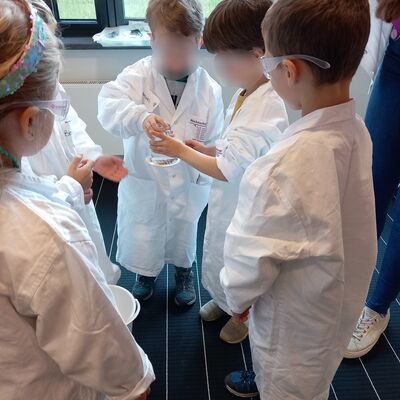 Kinder in wei&szlig;en Kitteln und mit Schutzbrillen stehen im Kreis und schauen auf ein Glas, dass eines der Kinder in der Hand h&auml;lt.
